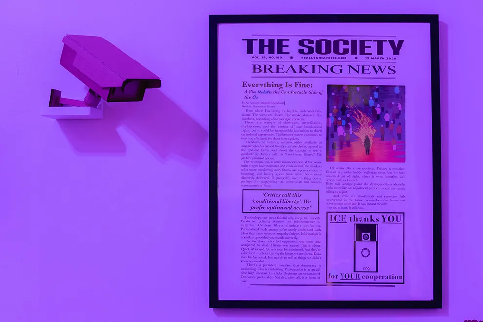 Purple-lit gallery wall featuring a framed fictional newspaper front page titled “THE SOCIETY,” alongside a mounted surveillance camera, evoking themes of surveillance, control, and media influence.